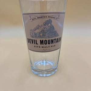 May include: A clear pint glass with a tan label featuring the text "Devil Mountain Brewing Co." and an illustration of a train. The glass also has the text "Five Malt Ale."