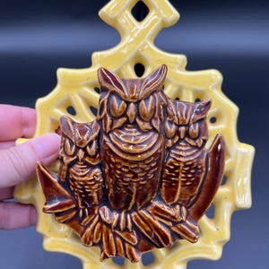 May include: A ceramic wall hanging featuring three brown owls perched on a branch, set against a yellow, openwork background. The owls have detailed feathers and expressive eyes. The piece has a glossy finish and a decorative top.