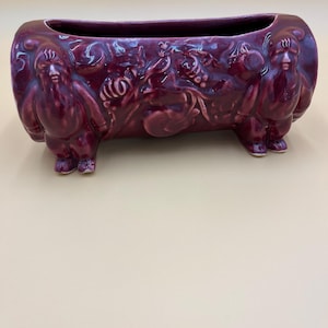 May include: A burgundy ceramic planter with a rectangular shape and a detailed relief design. The planter features figures on each end and a central relief of foliage and faces. The planter is raised on small feet.