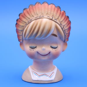 May include: A vintage ceramic figurine of a smiling child with closed eyes, wearing a headdress with pink and tan feathers. The figurine has a tan face with rosy cheeks and a white bib with a red and blue zigzag design. The background is a solid blue.
