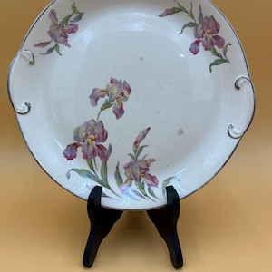 May include: A white ceramic serving plate with a silver rim and two handles. The plate is decorated with purple and yellow iris flowers and green leaves. The plate is displayed on a black stand.