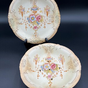 May include: Two antique porcelain plates with a floral design. The top plate is plain, while the bottom plate has holes in the center. Both plates have a cream background with gold accents.