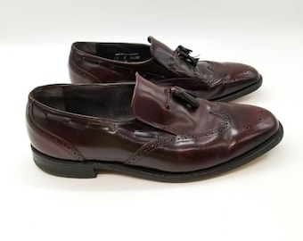 May include: A pair of burgundy leather tassel loafers with a brogue design. The shoes have a black leather sole and a tassel detail on the vamp.
