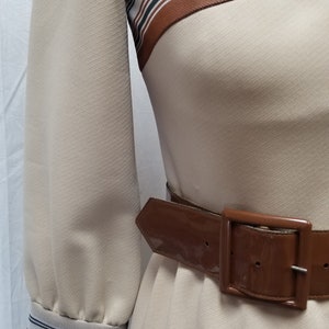 May include: A beige dress with brown and white striped trim at the neckline and cuffs. The dress has a brown patent leather belt with a silver buckle.