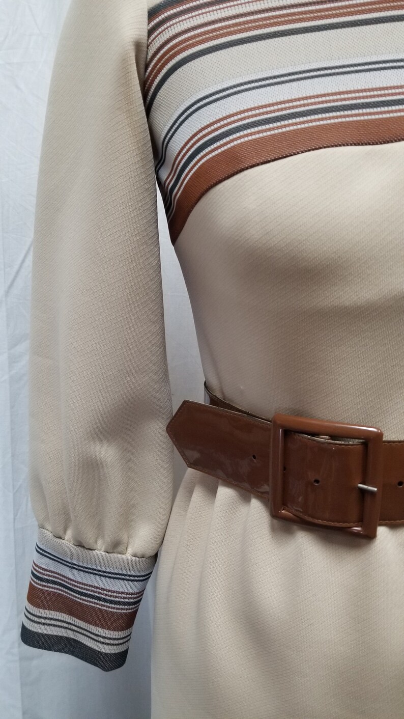 May include: A beige dress with brown and white striped trim at the neckline and cuffs. The dress has a brown patent leather belt with a silver buckle.