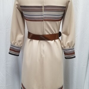 May include: A beige dress with brown and white striped bands around the neckline, waist, and hem. The dress has a brown belt and a zipper down the back.