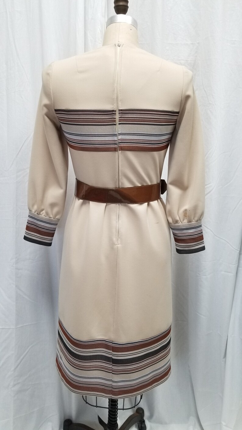 May include: A beige dress with brown and white striped bands around the neckline, waist, and hem. The dress has a brown belt and a zipper down the back.