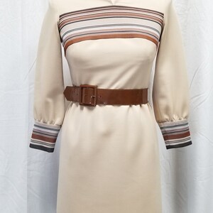 May include: A beige dress with brown and black striped trim at the neckline, cuffs, and hem. The dress has a brown patent leather belt.