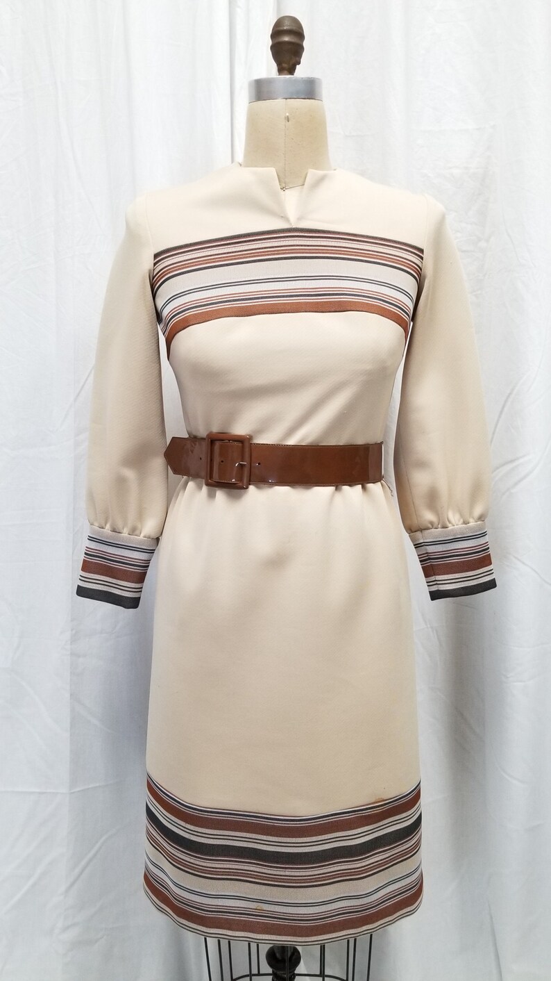 May include: A beige dress with brown and black striped trim at the neckline, cuffs, and hem. The dress has a brown patent leather belt.