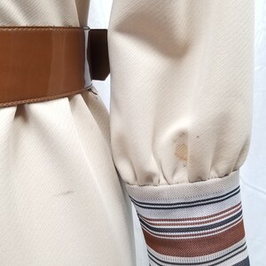 May include: A beige dress with a brown belt and striped cuffs. The dress has a textured fabric and a slight sheen.