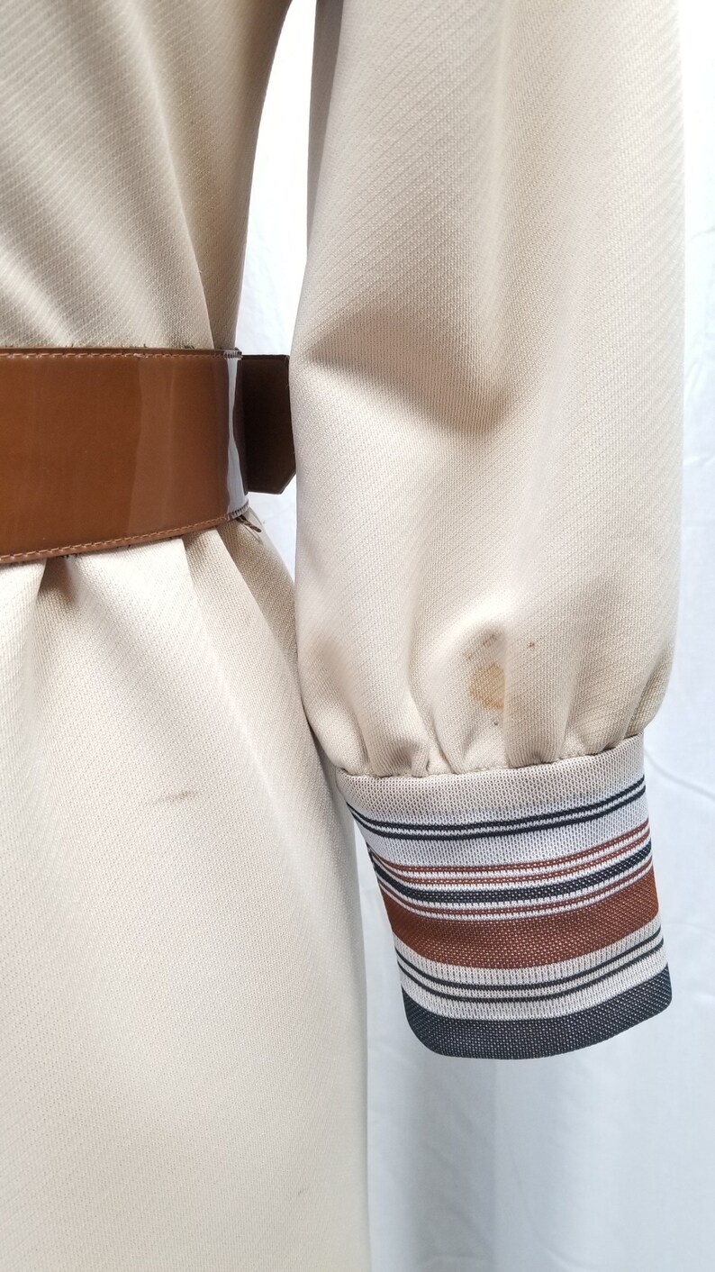 May include: A beige dress with a brown belt and striped cuffs. The dress has a textured fabric and a slight sheen.