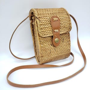 Könnte beinhalten: Eine rechteckige Umhängetasche aus geflochtenem Rattan mit Klappenverschluss und braunem Lederriemen. Die Tasche hat einen braunen Lederriemen und einen Knopfverschluss. Die Tasche ist zum Über-der-Schulter-Tragen konzipiert.