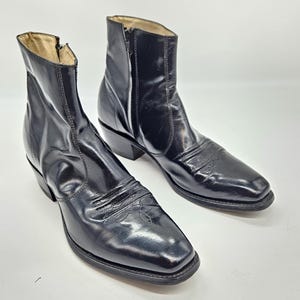 Puede incluir: Un par de botines de cuero negro con punta cuadrada y tacón bajo. Las botas tienen una cremallera lateral y costuras decorativas en la pala. El forro interior es de color beige claro.