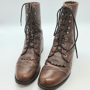 Op de afbeelding: Een paar bruine leren veterlaarzen. De laarzen hebben een getextureerde uitstraling, een franjedetail op de neus en metalen veterogen. De veters zijn donkerbruin.