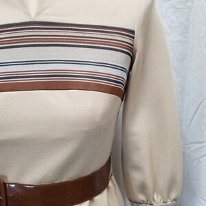 May include: A beige dress with brown and white striped detail at the neckline and cuffs. The dress has a brown belt.