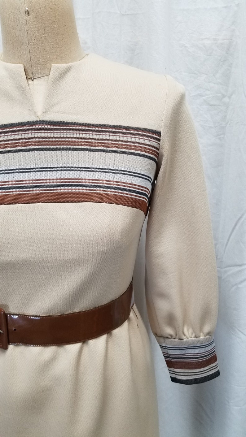 May include: A beige dress with brown and white striped detail at the neckline and cuffs. The dress has a brown belt.