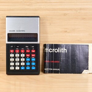 May include: A vintage Microlith scientific calculator with a silver top and black base, featuring red, blue, and white keys. Next to it is a black instruction manual with the brand name "microlith" in white.