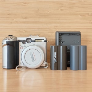 May include: A silver and black Canon PowerShot G6 digital camera with a lens cap, two batteries, and a charger. The lens cap has the Canon logo. The batteries are labeled "maxell."