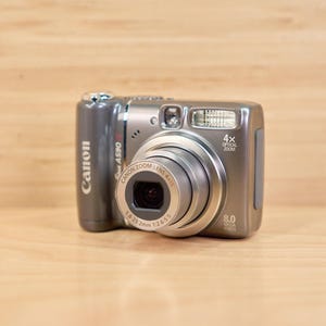 May include: A silver Canon PowerShot A590 IS digital camera. The camera has a 4x optical zoom lens and an 8.0-megapixel resolution. The camera is on a wooden surface.
