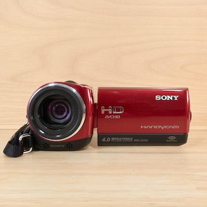 Pode incluir: Uma câmara de vídeo digital Sony Handycam vermelho-escura com uma lente proeminente, uma correia de pulso preta e um painel lateral com as etiquetas HD, AVCHD, 4.0 Mega Pixels e HDR-CX100. O dispositivo tem um acabamento brilhante e compacto.
