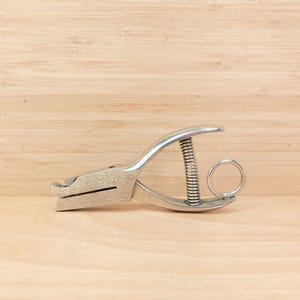 Op de afbeelding: Een zilverkleurige, handmatige perforator met een veer en een ronde handgreep. De tool is ontworpen om gaten in papier te maken. De achtergrond is een licht houten oppervlak.
