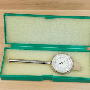 May include: A silver and white circular gauge with a black needle and a long, silver handle, inside an open, green rectangular case with a light yellow foam interior. The case is on a wooden surface.