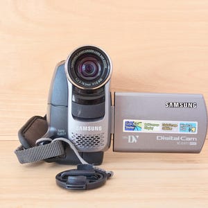 May include: A grey Samsung MiniDV digital camcorder with a flip-out LCD screen. The lens barrel features text reading Samsung Camcorder Lens 345x Zoom. The side panel displays the Samsung logo, Digital Cam SC-D371 text, and several small feature stickers.