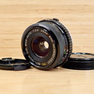 Könnte beinhalten: Ein schwarzes Soligor MC Wide-Auto 28mm Kameraobjektiv mit 49mm Durchmesser und einer Blende von 1:2.8. Das Objektiv hat einen schwarzen Objektivdeckel und einen hinteren Objektivdeckel. Auf einer Holzoberfläche.