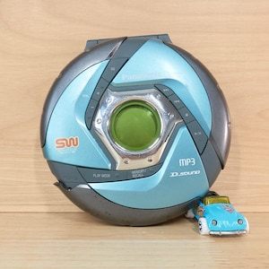 May include: A circular Panasonic portable CD player in metallic blue and dark gray with a central green circular window, featuring MP3 and D.SOUND branding, accompanied by a small light blue toy car with white wheels positioned at the bottom right.