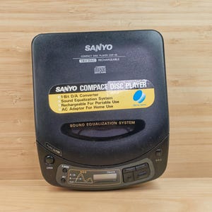 Op de afbeelding: Een zwarte Sanyo Compact Disc Player met een gele en blauwe sticker. De speler heeft een digitaal display en bedieningsknoppen. De tekst op de speler bevat "Compact Disc Player" en "Sound Equalization System".
