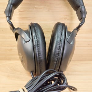 May include: Black over-ear headphones with a padded headband and earcups. The earcups are oval-shaped and have a leather-like texture. A black cable extends from the headphones. The background is a light brown wooden surface.