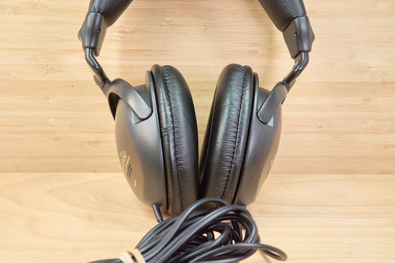 May include: Black over-ear headphones with a padded headband and earcups. The earcups are oval-shaped and have a leather-like texture. A black cable extends from the headphones. The background is a light brown wooden surface.
