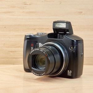 May include: A black Canon digital camera with a pop-up flash and a zoom lens. The lens has the text "Canon Zoom Lens 10x" and measurements of 6.0-60.0mm 1:2.8-4.3. The camera is on a light wood surface.