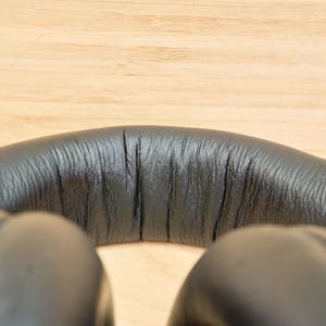 May include: Close-up of black over-ear headphones. The headband is made of textured black material, with visible stitching and a slight curve. The earcups are also black and visible at the bottom of the frame.
