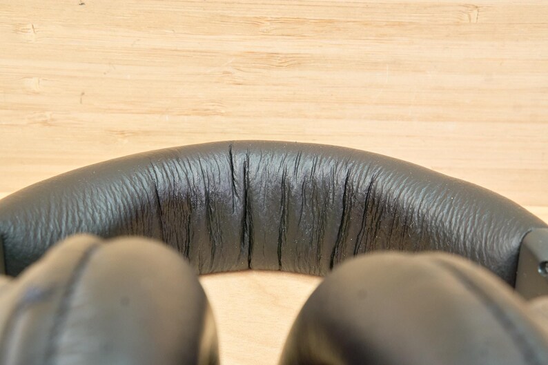 May include: Close-up of black over-ear headphones. The headband is made of textured black material, with visible stitching and a slight curve. The earcups are also black and visible at the bottom of the frame.
