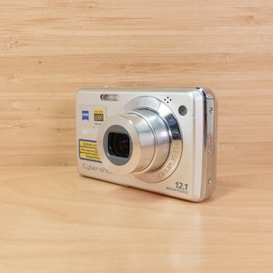 May include: Silver digital camera with a prominent lens and the text "Cyber-shot" visible. The camera has a 12.1 megapixel resolution and features a Zeiss lens. The camera is on a wooden surface.