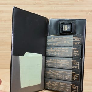 Op de afbeelding: Zwarte leren etui met een Master Library Module en verschillende Solid State Software kaarten. De kaarten zijn gelabeld met wiskundige functies en vergelijkingen. Een kleine stapel lichtgroen papier is zichtbaar in de etui.