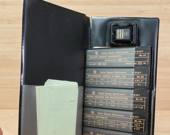 Può includere: Custodia in pelle nera con un modulo Master Library e diverse schede Solid State Software. Le schede sono etichettate con funzioni ed equazioni matematiche. Una piccola pila di carta verde chiaro è visibile all'interno.