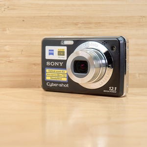 Könnte beinhalten: Schwarze Sony Cyber-shot Digitalkamera mit silbernem Objektiv und 12,1 Megapixel Auflösung. Funktionen: optischer Zoom, Weitwinkel und SteadyShot. Die Kamera liegt auf einer hellbraunen Holzoberfläche.