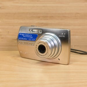 以下が含まれることがあります： 7.1メガピクセルのレンズを搭載したシルバーのデジタルカメラ。カメラには、「Bright Capture Technology」と「All-Weather 2.5" LCD」と書かれた青いラベルが付いています。