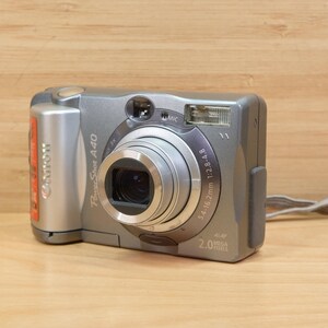 May include: Silver Canon PowerShot A40 digital camera with a retractable lens and a built-in flash. The camera has a silver body with a black lens and a small hand strap. The camera is on a wooden surface.