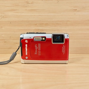 May include: A red and silver Olympus Tough digital camera with a grey wrist strap. The camera has the word "Tough" printed on the front. The camera is waterproof and shockproof.