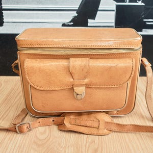 May include: A vintage tan leather camera bag. It has a rectangular shape, a front pocket, and a shoulder strap. The bag features a zip closure and a metal clasp on the front pocket. It is on a wooden surface.