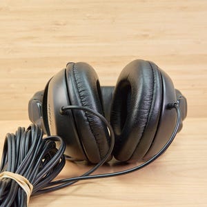 May include: Black over-ear headphones with a coiled black cable secured with a rubber band. The headphones have large, padded earcups and a connecting headband. The background is a light brown wooden surface.