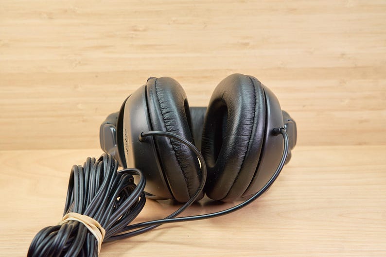 May include: Black over-ear headphones with a coiled black cable secured with a rubber band. The headphones have large, padded earcups and a connecting headband. The background is a light brown wooden surface.