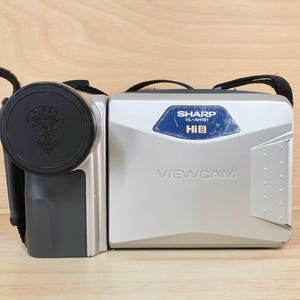 Op de afbeelding: Een zilverkleurige Sharp Viewcam VL-AH151 Hi8 camcorder met een zwarte lensdop aan de linkerkant. Het apparaat heeft een rechthoekige behuizing met een blauw label waarop de merknaam, het modelnummer en de Hi8-tekst staan, plus een zwarte draagriem.