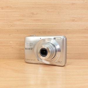 May include: Silver Sony Cyber-shot digital camera with a prominent lens and flash. The camera has the text "SONY Cyber-shot 14.1 MEGA PIXELS" printed on the front. The camera is on a light brown wooden surface.