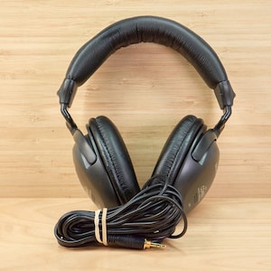 May include: Black over-ear headphones with a padded headband and earcups. The headphones have a coiled black cord with a 3.5mm audio jack. The cord is secured with a rubber band. The headphones are on a light brown wooden surface.