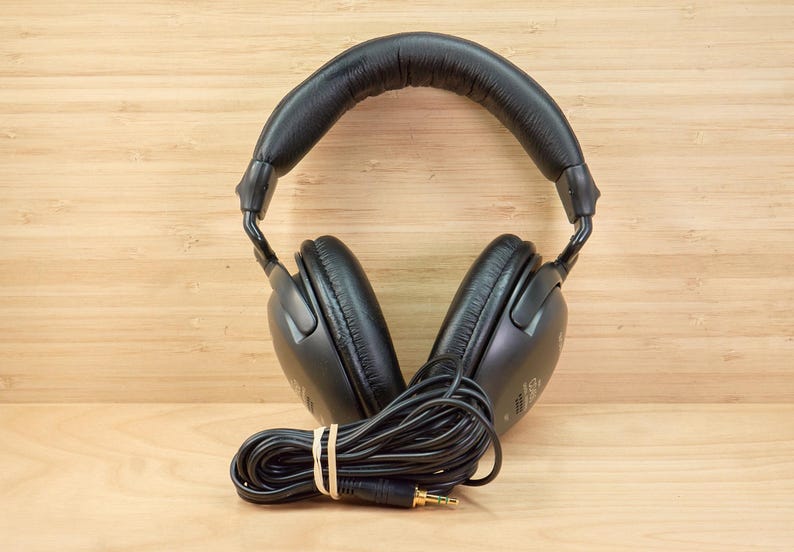 May include: Black over-ear headphones with a padded headband and earcups. The headphones have a coiled black cord with a 3.5mm audio jack. The cord is secured with a rubber band. The headphones are on a light brown wooden surface.
