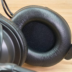 May include: Close-up of black over-ear headphones. The earcups are made of a soft, textured material, with a circular opening covered by a black, porous fabric. The headphones have a padded headband and are designed for comfort and sound isolation.